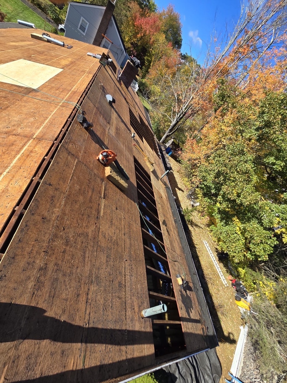 Aerial drone view of a roof mid-tear-off, autumn tree line, ladder against the eave