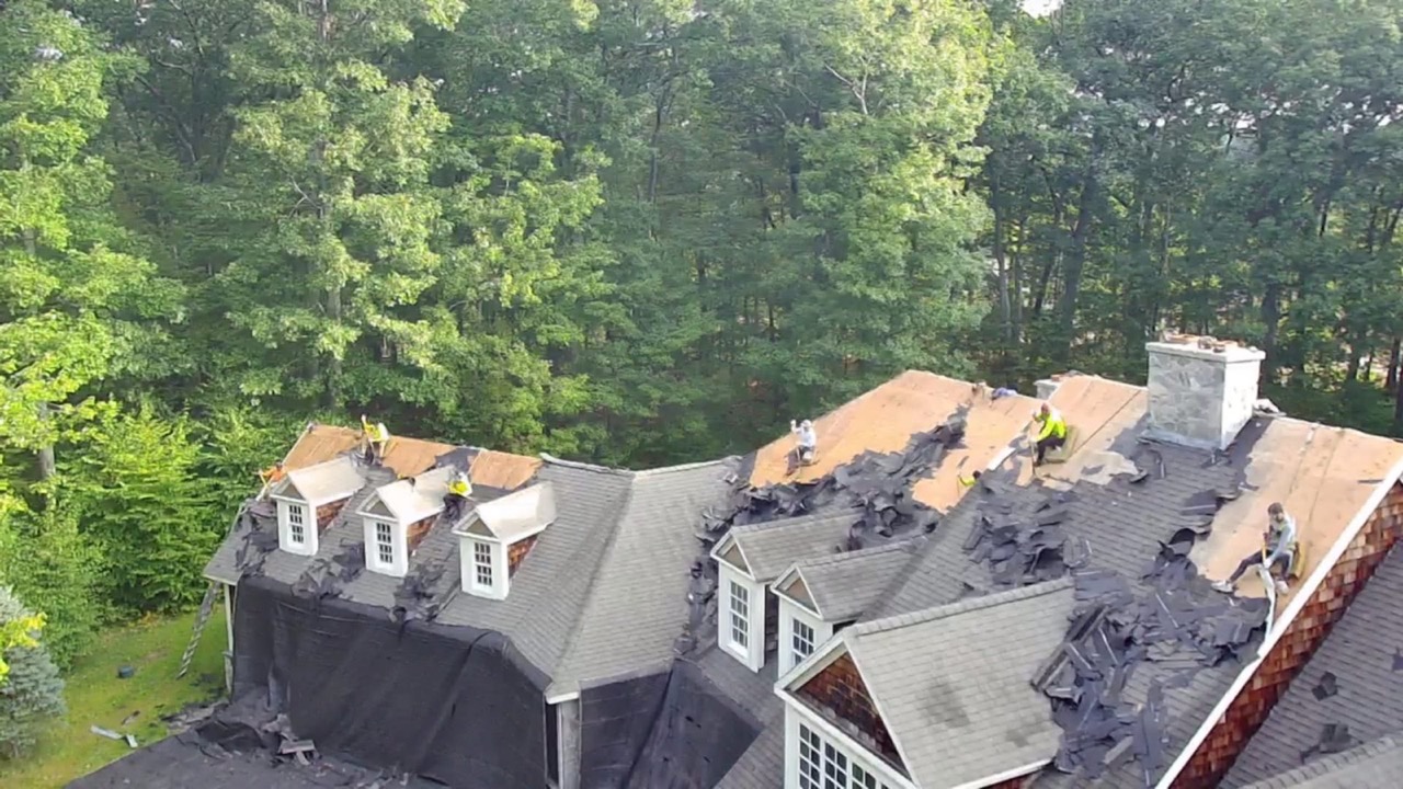 Aerial drone photo of the Genesis General Construction crew mid-tear-off on a large multi-dormer home, decking exposed, working in the tree line above Waterbury, CT